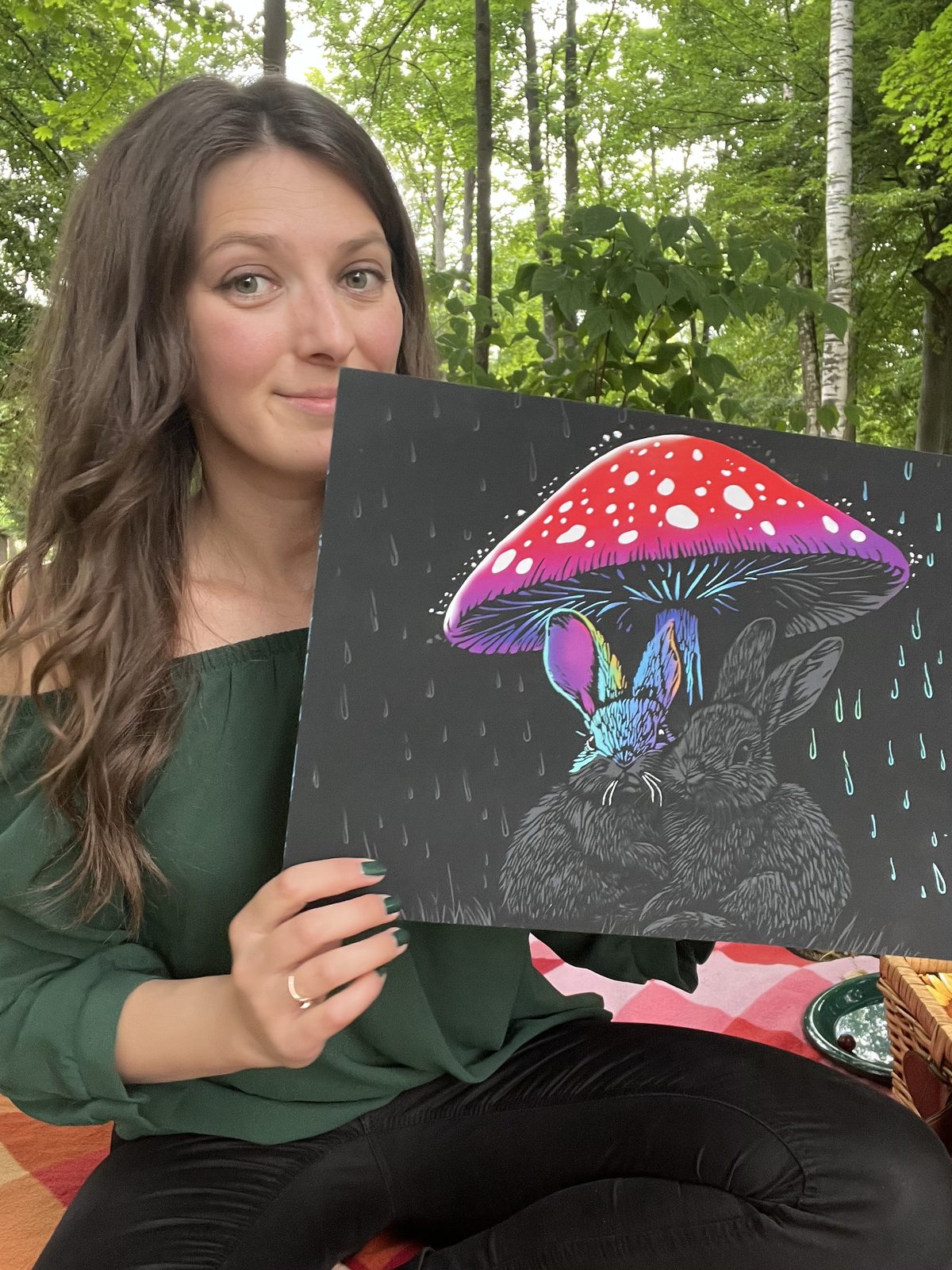 Smiling woman holding finished mushroom-and-rabbit canvas in a forest